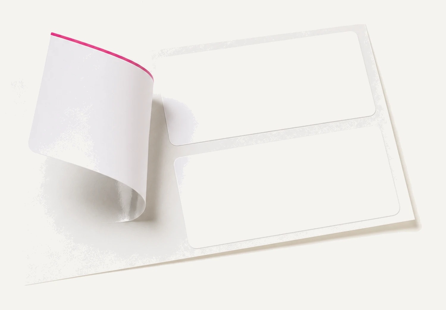 One blank white adhesive label peeled halfway off its backing sheet and curling upward, two unpeeled labels still beside it, the lifted label trimmed in magenta.