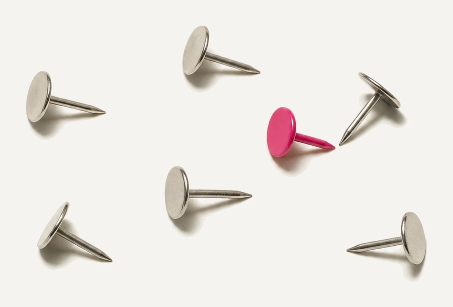 Seven small steel push pins scattered loosely across the surface at different angles, each casting a small sharp shadow, one of them with a magenta head.