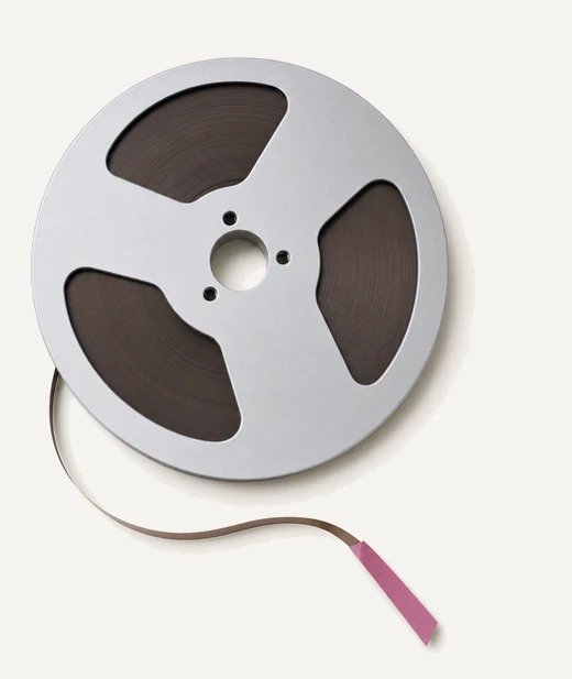 A reel of quarter inch magnetic tape on a metal spool, a short tail of tape curving away with a magenta tab clipped to it.